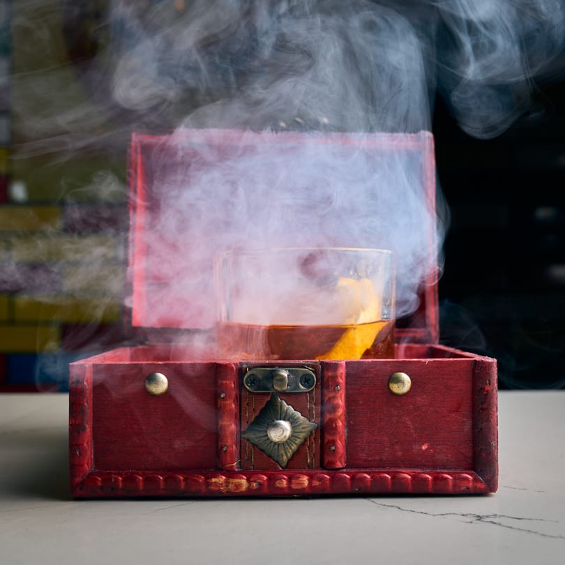 Smoked cocktail in a glass with an orange peel, presented in an open red wooden box.