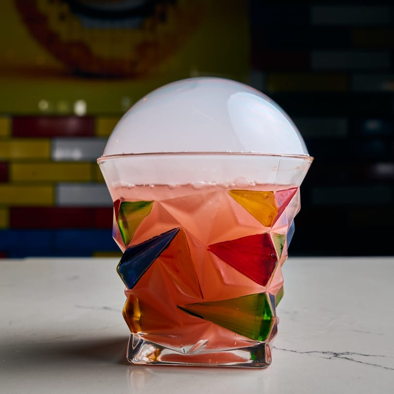 A vibrant, colorful cocktail in a geometric glass, topped with a delicate smoke bubble, on a white marble surface.